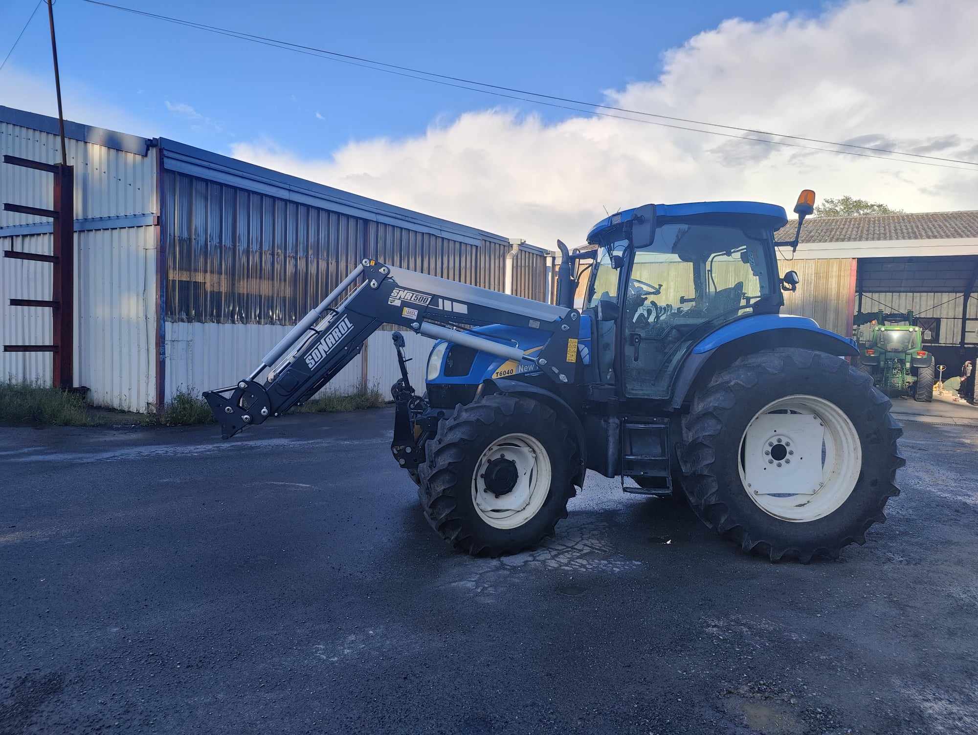 Tracteur bleu avec chargeur frontal Stoll Solid devant hangars agricoles md-services-mecanique-hydraulique-aveyron en Aveyron